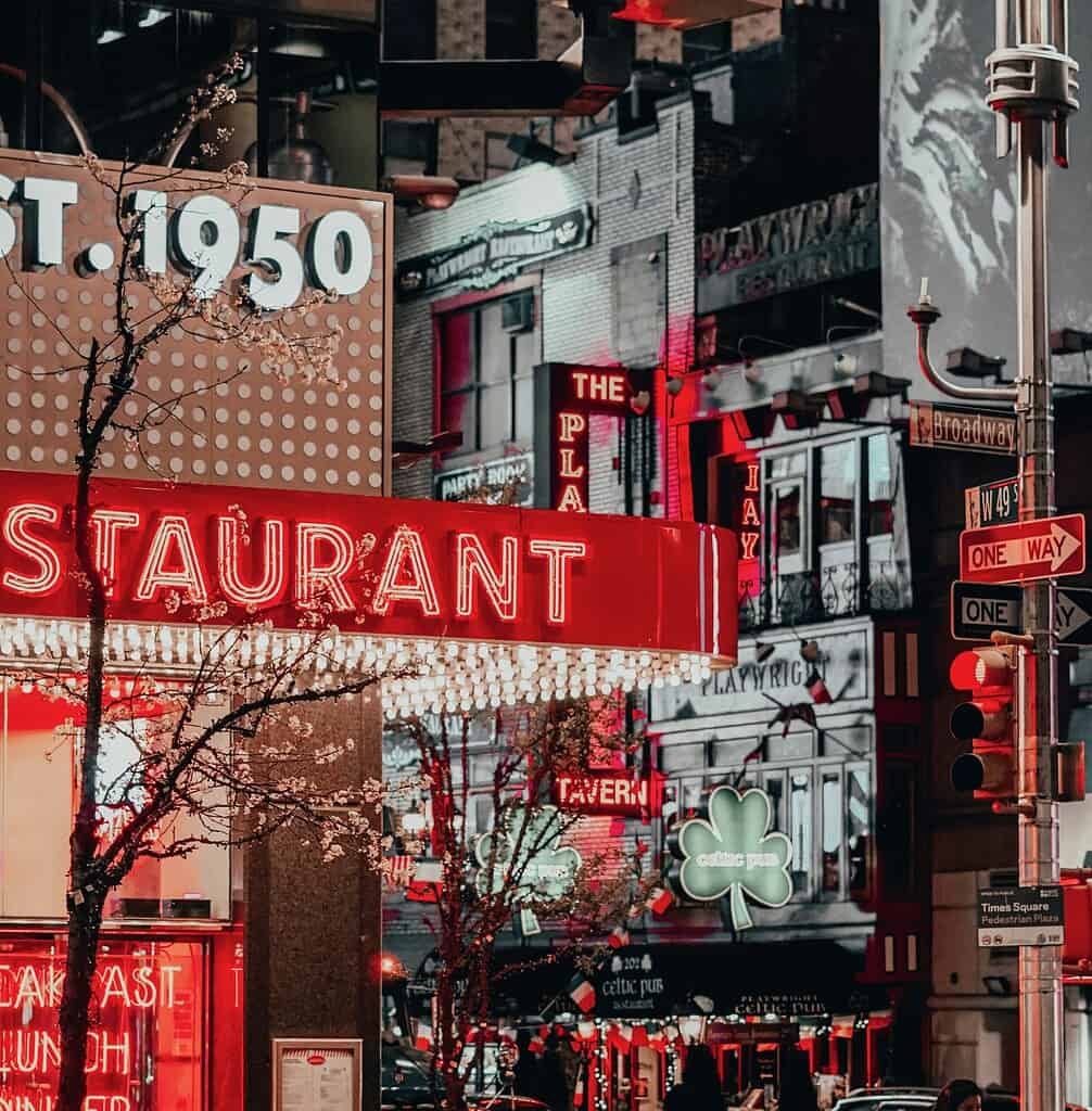 A brightly lit restaurant exterior in Times Square at night, featuring glowing neon signs, massive digital billboards in the background, and the vibrant energy of the city lights.