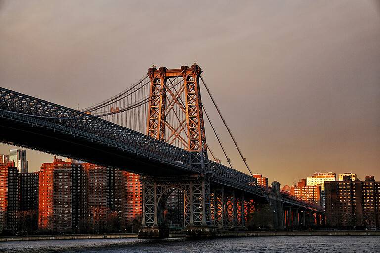 A golden sunset over the Williamsburg Bridge in the summer, with the sun low in the sky casting a warm glow over the bridge's steel suspension towers and the East River.